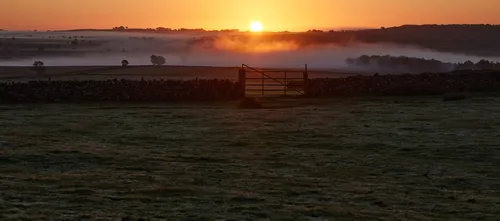 This landscape photograph captures a misty gate leading through a rural field in the Peak District near Curbar Edge at sunrise. The early morning light of autumn creates a soft orange glow across the horizon, illuminating layers of mist that drift above the undulating countryside. A dry stone wall runs across the foreground, typical of rural boundaries in this region, and adds texture to the scene. The sun rises behind distant hills, producing long shadows and enhancing the atmospheric mist over the landscape. The combination of the gate, wall, mist, and low sun signifies the tranquil beauty of an autumn morning in the Peak District.